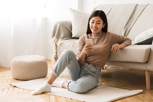 Happy Asian Girl Messaging On Phone At Home, Sitting On Floor Near Sofa